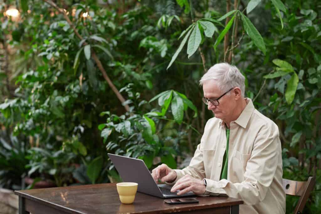 older man working on a laptop outdoors surrounded by plants showing stock vest during notice period